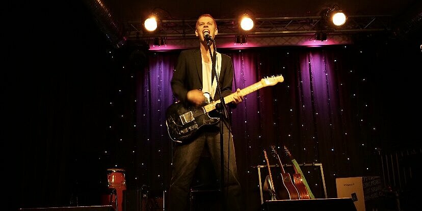 Ein Mann steht mit E-Gitarre an einem Mikrofon.