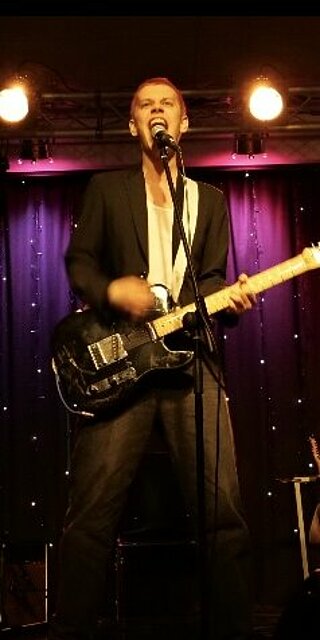Ein Mann steht mit E-Gitarre an einem Mikrofon.