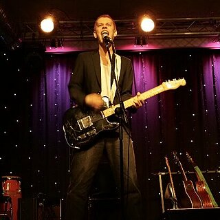 Ein Mann steht mit E-Gitarre an einem Mikrofon.