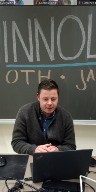Ein Mann steht in einem Vorlesungsraum vor einer Tafel. Der Vortrag wird auch virtuell übertragen.